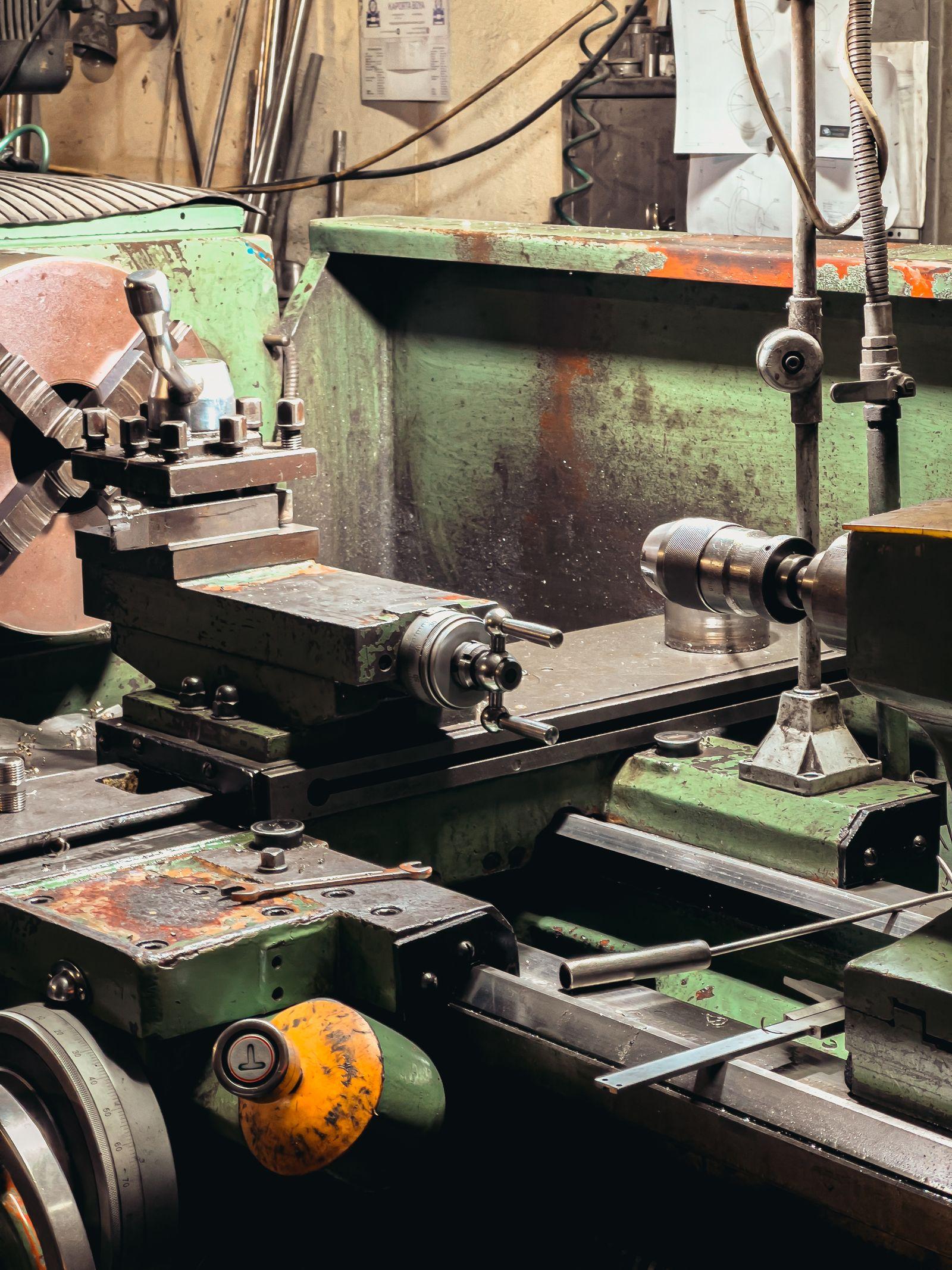 Manual lathe with chuck and toolpost, engineering drawing pinned on the wall behind
